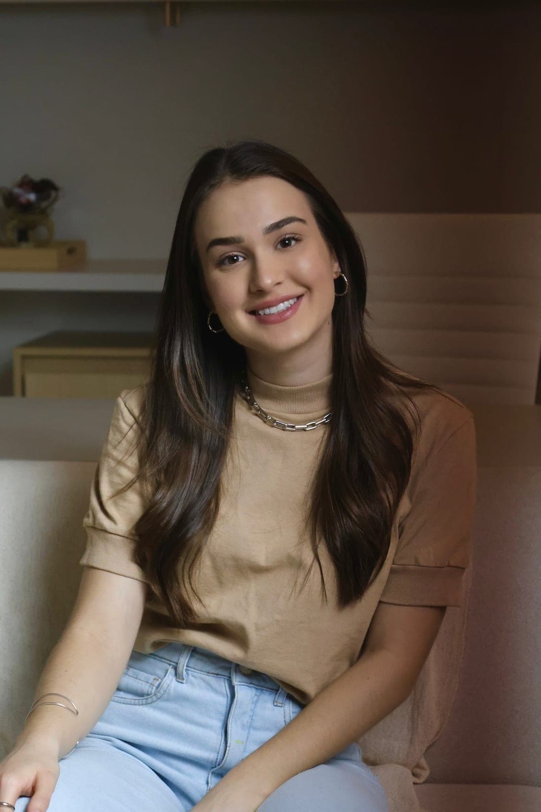 Psicóloga Ana Carolina Crema sentada no consultório, de blusa bege