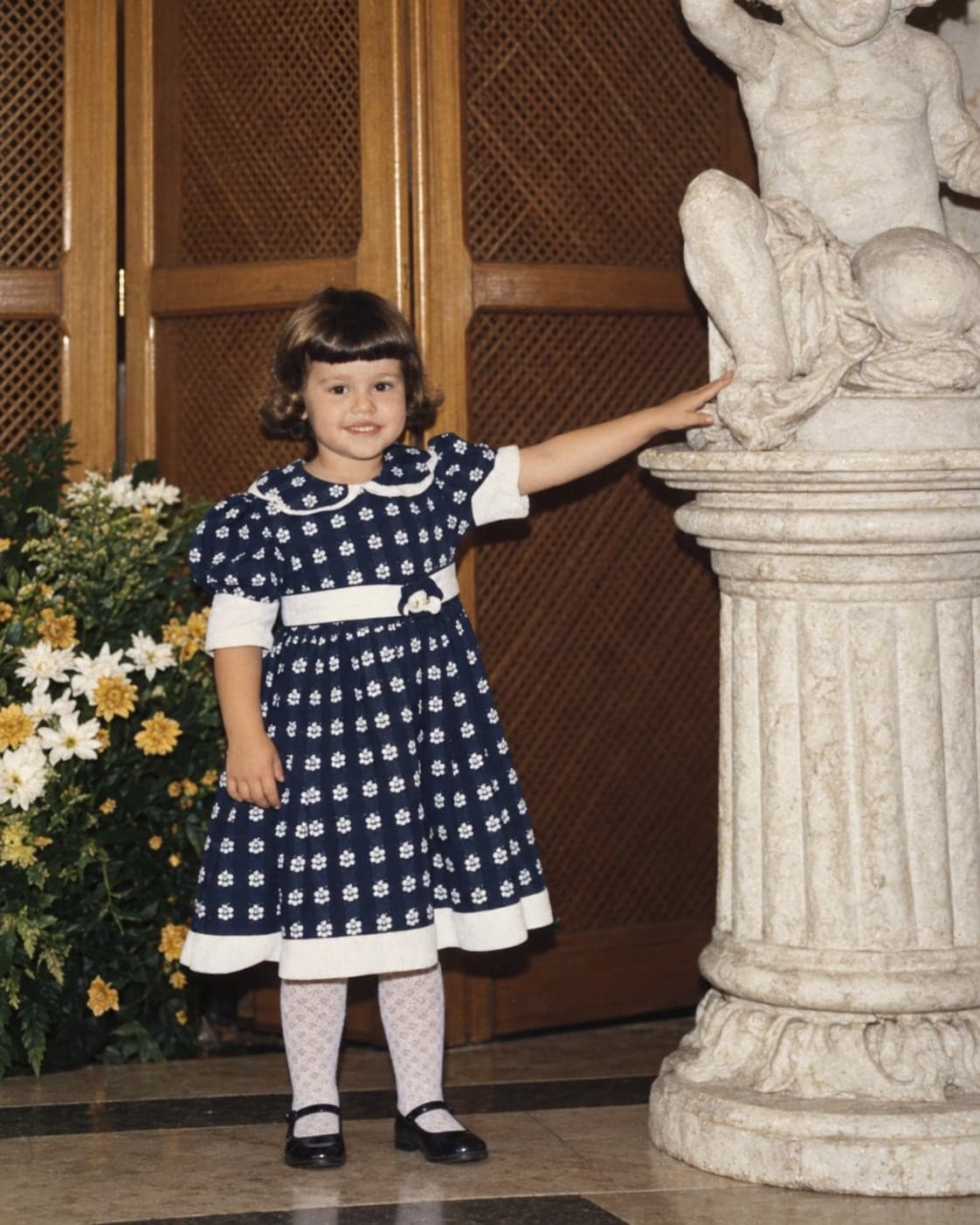 Carol criança, de vestido azul, ao lado de uma estátua e flores